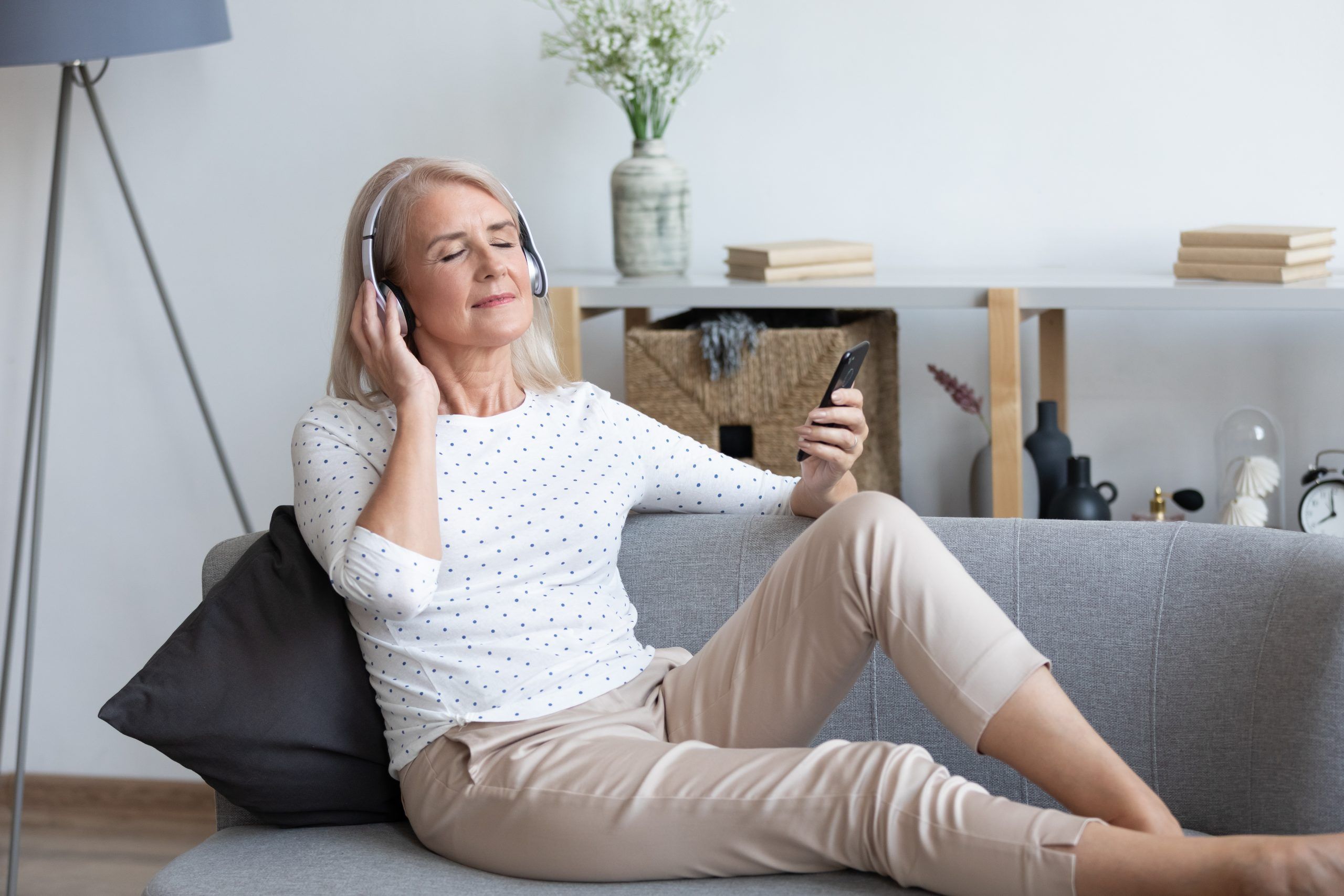 Full length peaceful mature retired lady enjoying favorite music in earphones, chilling on comfortable sofa at home. Tranquil older woman listening to classic tracks, chosen from mobile application.