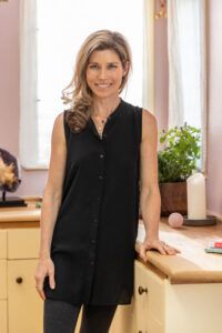 Smiling woman with light brown hair in a black sleeveless button-down top standing in a bright kitchen, hand resting on a counter.
